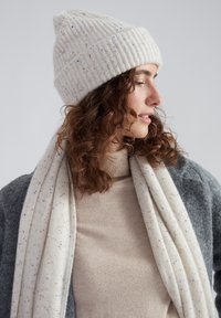 Brede gebreide muts in crèmekleur met spikkels, gecombineerd met een zachte sjaal met een vergelijkbaar patroon. Het model draagt een beige coltrui en een grijze cardigan.