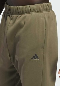 Olivegrønne basketball-sweatpants med elastisk talje, sidelommer og et sort Adidas-logo med teksten "BASKETBALL". Glat tekstur.