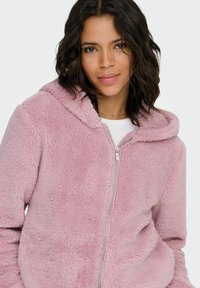 Roze fleece hoodie met een rits aan de voorkant en een gevoerde capuchon. Heeft een zachte, gestructureerde oppervlakte en zijzakken. Effen wit aan de binnenkant.