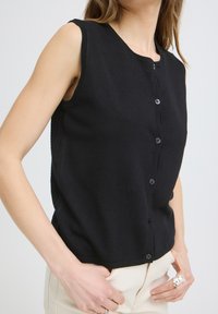 Femme portant un gilet noir sans manches boutonné et un pantalon beige clair, avec des bagues en argent aux doigts de la main droite.