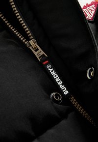 Crna puffer jakna s brončanim zatvaračem, koja sadrži istaknutu oznaku "Superdry" i detalje od dugmadi. Glatka tkanina.