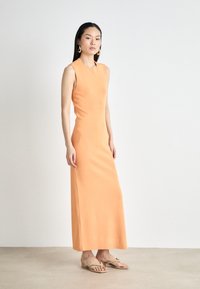 Maxikjole i blødt orange stof, ærmeløs design, tætsiddende silhuet, glat tekstur, suppleret med beige flip-flops for en afslappet stil.