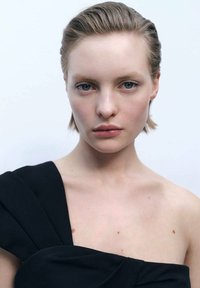 Black one-shoulder dress with draped fabric, smooth texture, and minimalist design. Model's face has natural makeup and short, slicked-back hair.
