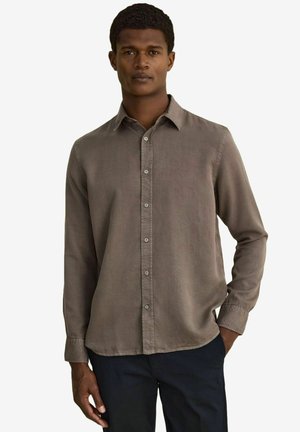 Man som bär en brun långärmad skjorta med knappar och mörka byxor, stående med ena handen delvis i fickan mot en enfärgad bakgrund.