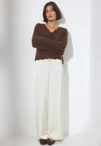 Bruine, gebreide trui met een scalloped halslijn, gecombineerd met witte, wijde satijnen broek en zwarte, puntige schoenen.