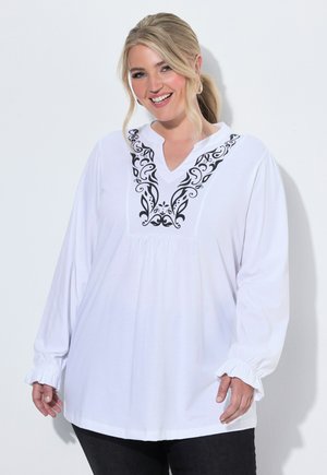 Lächelnde Frau mit blonden Haaren trägt eine weiße Bluse mit langen Ärmeln und schwarzen dekorativen Mustern auf der Brust sowie schwarze Hose.