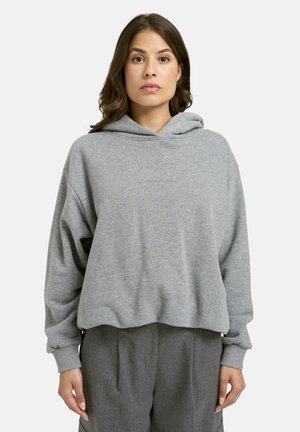 Woman with shoulder-length brown hair wears a loose gray hoodie and gray pants, standing against a plain light background.