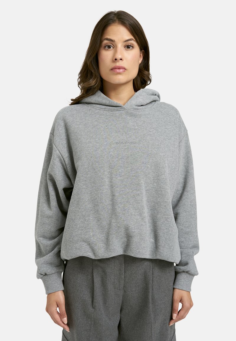 Frau mit schulterlangem braunem Haar trägt einen lässigen grauen Hoodie und graue Hosen und steht vor einem schlichten hellen Hintergrund.