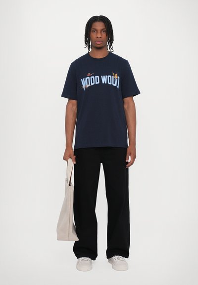 Camiseta de algodón azul marino con estampado "WOOD WOOD" en azul claro, combinada con pantalones de pierna ancha negros y una bolsa tote beige. Incluye calzado casual.