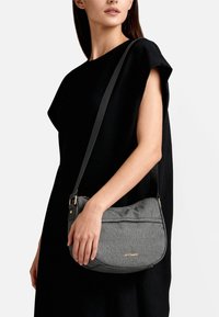 Borsa a tracolla grigia con texture e forma curva, tracolla nera, logo color oro e chiusura con cerniera, tenuta contro un indumento nero.