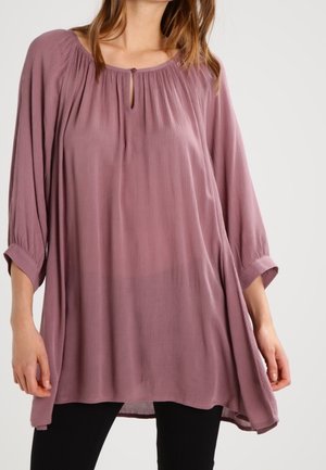 Kvinde iført en løs, langærmet mauve bluse med knapper ved nøglehulshals og sorte bukser mod en ensfarvet baggrund.