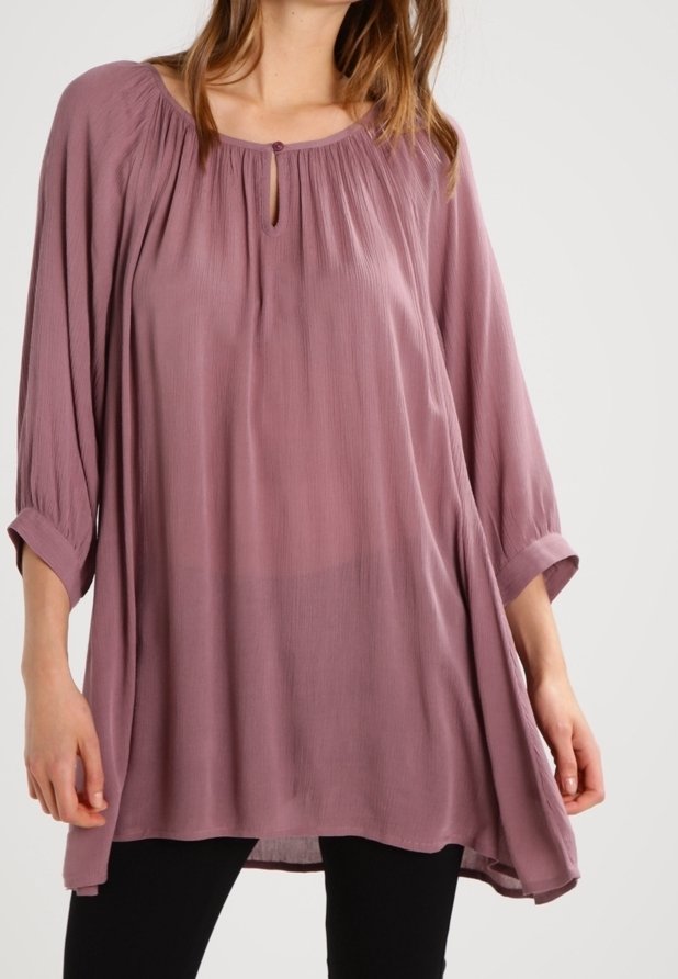 Femme portant un chemisier ample mauve à manches longues avec un col en forme de trou de serrure boutonné et un pantalon noir sur un fond uni.