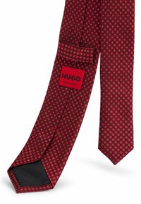 Röd silkesnära med diamantmönster och silverdetaljer. Innehåller en etikett med texten "HUGO MADE IN ITALY." Slät textur, smal design.