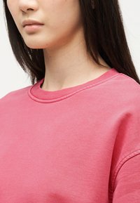 Gros plan d'une personne portant un sweat-shirt rose uni à col rond, montrant l'encolure, l'épaule et le bas du visage.