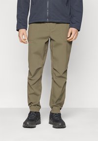 Calças caqui com um acabamento texturizado, apresentando bolsos laterais e punhos elásticos, combinadas com sapatos de caminhada pretos e uma jaqueta azul-marinho.
