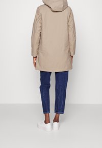 Beige parka met een capuchon, met een rechte snit, lange mouwen en een gladde textuur. Gecombineerd met donkerblauwe jeans en witte sneakers.