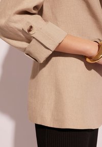 Beżowa lniana marynarka z podwiniętym mankietem i detalami na guziki, w zestawie z czarnymi prążkowanymi spodniami. Charakteryzuje się elegancką sylwetką i lekką teksturą.