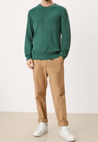Homme portant un pull vert côtelé, un pantalon chino beige retroussé, des chaussettes blanches et des baskets blanches, debout avec une main dans la poche sur un fond uni.