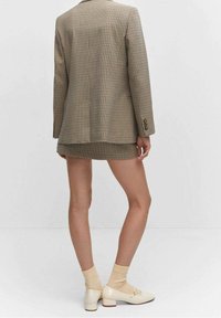 Blazer à carreaux marron avec une fente unique, associé à une mini-jupe à carreaux assortie. Des chaussettes claires et des chaussures crème à talons bas complètent la tenue.