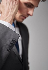 Veste de costume grise avec quatre boutons noirs sur le poignet, tissu texturé, col de chemise blanc visible et une cravate bleue. Gros plan sur une main ajustant la veste.