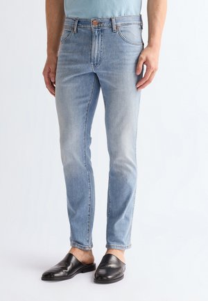 Homme portant un jean slim bleu clair et des chaussures en cuir noires sans lacets, debout devant un fond blanc.