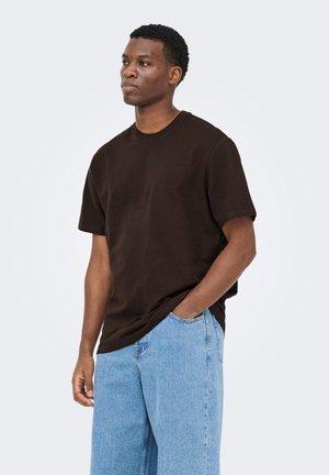 Jeune homme aux cheveux courts et bouclés portant une chemise marron foncé à manches courtes et un jean bleu clair, debout avec une main dans la poche.