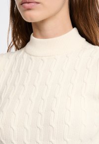 Zbliżenie na osobę noszącą sweter w kolorze kremowym z warkoczowym splotem i żebrowanym kołnierzem typu mock neck.