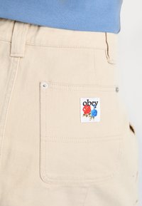 Beiga byxor har en fyrkantig bakficka med en texturerad etikett som visar ordet "obey" och blommiga accenter i rött och blått.