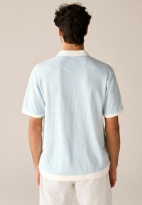 Vaaleansininen lyhythihainen poolopaita, jossa on teksturoitu kuvio, valkoinen kaulus ja kalvosimet, pehmeästä kankaasta, esitetty takaa.