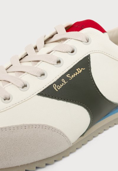 Basket blanche avec bout en daim beige, panneau latéral vert foncé, languette rouge au talon, lacets beige et "Paul Smith" écrit en or sur le côté.