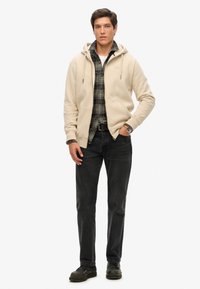Sudadera beige con cremallera sobre una camisa de cuadros, jeans oscuros y zapatos negros. La sudadera tiene cordones; la camisa presenta un patrón de cuadros multicolor.
