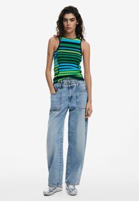 Canottiera senza maniche a righe in verde, blu e nero; abbinata a jeans a gamba larga blu chiaro. Modella con le mani nelle tasche. Sneakers argentate.