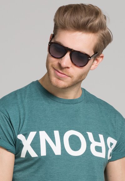 Roheline T-särk, millel on valge peatähega tekst "BRONX". Mudel kannab tumedaid päikeseprille kilpkonnakuju raamiga ning heledate aktsentidega. Sile kangastekstuurne.