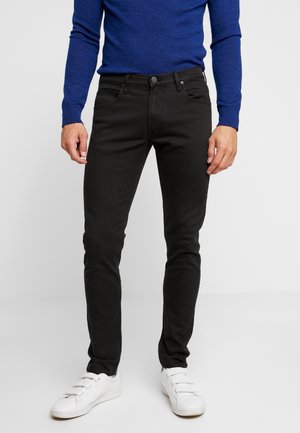 Homme portant un jean noir slim, des baskets blanches et un haut bleu à manches longues, debout sur un fond clair uni.