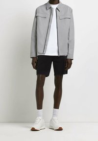 Giacca zip grigia chiara con due tasche sul petto, t-shirt bianca, pantaloni corti neri e scarpe da ginnastica bianche con dettagli marroni.