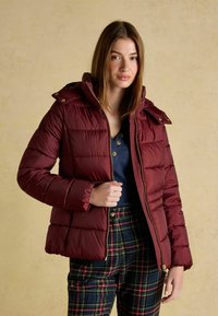 Burgundy puffer jacket with a zip front, attached hood, and ribbed cuffs. Paired with plaid trousers in dark green, red, and yellow tones.