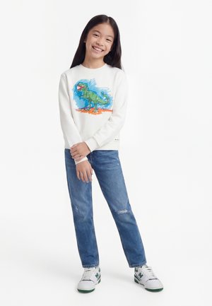 Fille souriante debout, portant un sweat-shirt blanc avec un motif de dinosaure vert, un jean bleu avec de petites déchirures, et des baskets blanches avec des détails verts.