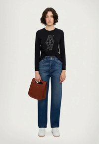 Sieviete ar īsiem, cirtainiem matiem, ģērbta melnā garo pied kuru krekla ar "AX" logotipu, zilajās džinsās, baltās sporta kurpēs, labajā rokā tur brūnu rokassomu.