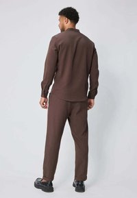 Chemise à manches longues marron et pantalon assorti en tissu lisse et texturé, avec une coupe décontractée et des poignets au niveau des poignets. Chaussures noires.