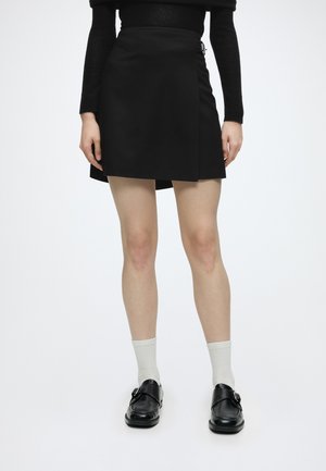 Person trägt einen schwarzen Minirock mit seitlicher Schnalle, ein schwarzes Langarmshirt, weiße Socken und schwarze Leder-Loafer und steht vor weißem Hintergrund.