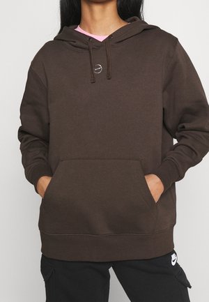Persona que lleva una sudadera con capucha marrón oscuro con bolsillo delantero y pantalones negros con el logo de Nike, con las manos en los bolsillos de la sudadera, sobre un fondo claro y liso.