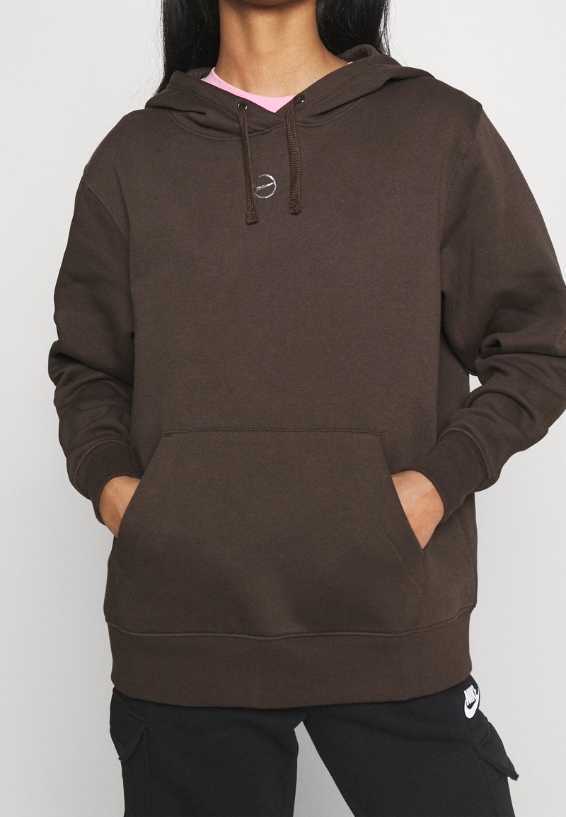 Personne portant un sweat à capuche marron foncé avec une poche avant et un pantalon noir avec le logo Nike, les mains dans les poches du sweat à capuche, sur un fond clair uni.