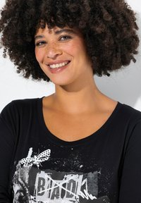 T-shirt noir à manches longues avec un imprimé graphique et des accents blancs, encolure ronde, et tissu doux avec un léger éclat. Cheveux bouclés visibles.