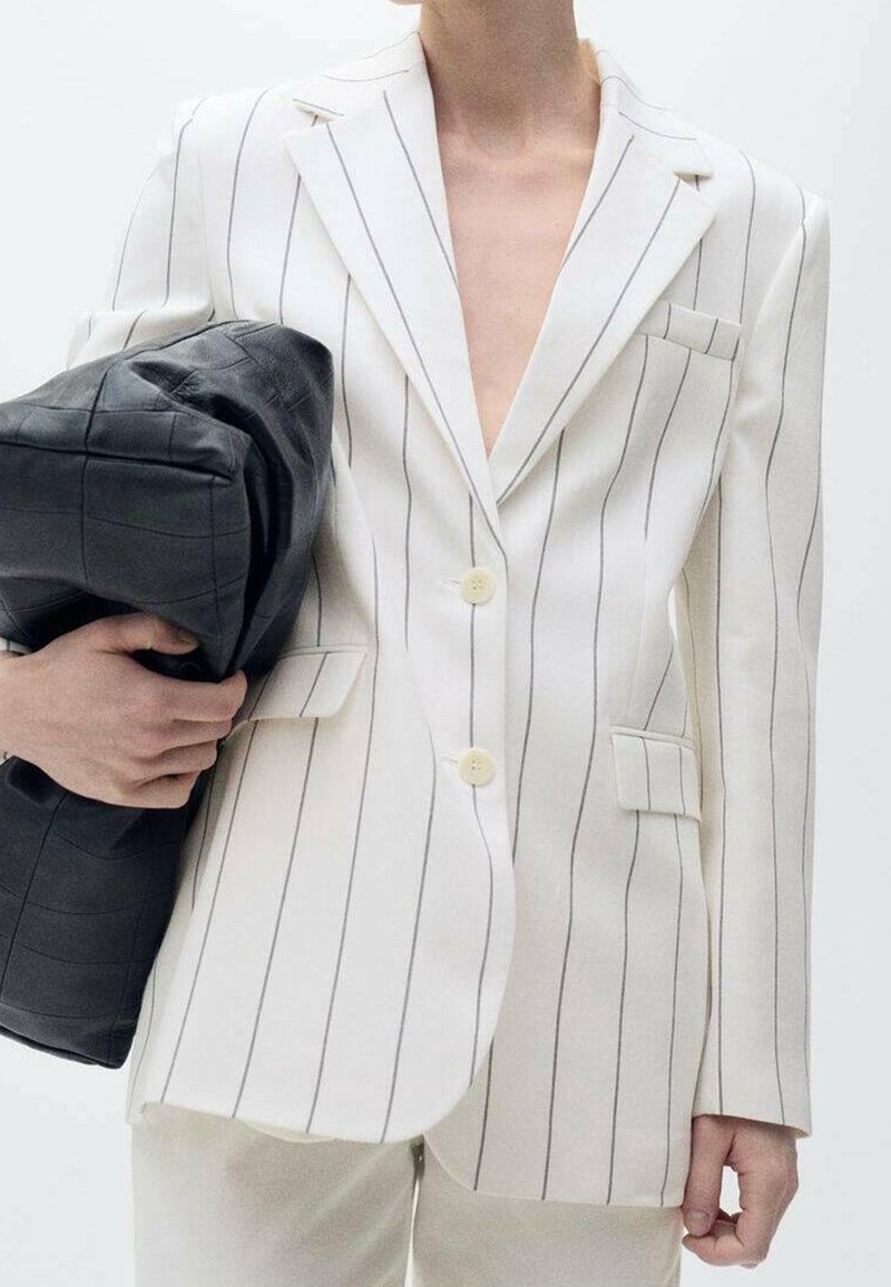 Blazer blanc à fines rayures avec deux boutons, poche avant et revers crantés. Tient un sac noir matelassé à la texture lisse.
