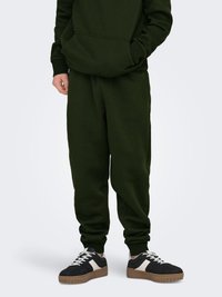 Sweat à capuche en coton vert foncé avec une poche avant, assorti à un pantalon de survêtement assorti. Baskets noires et blanches avec une semelle texturée.