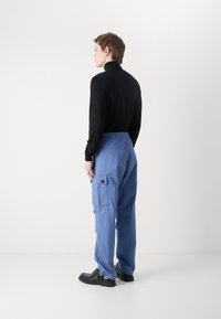 Calças de carga azuis com cintura elástica, bolsos laterais e detalhes em botão. Combinadas com um suéter preto justo, vistas pelas costas.