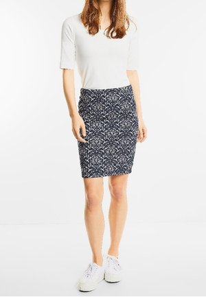 Vrouw draagt een wit kortärmelig shirt, een knielange marineblauwe rok met bloemenpatroon en witte sneakers, staande tegen een effen achtergrond.