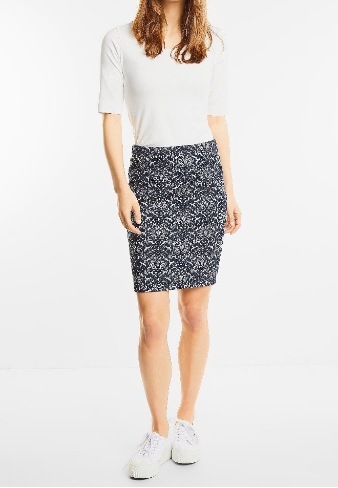 Femme portant un haut blanc à manches courtes, une jupe bleue marine à fleurs longueur genoux et des baskets blanches, debout devant un fond uni.