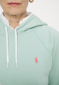 Mint green hoodie with white drawstrings, featuring a small pink embroidered logo on the left chest. Soft fabric with a classic fit.
