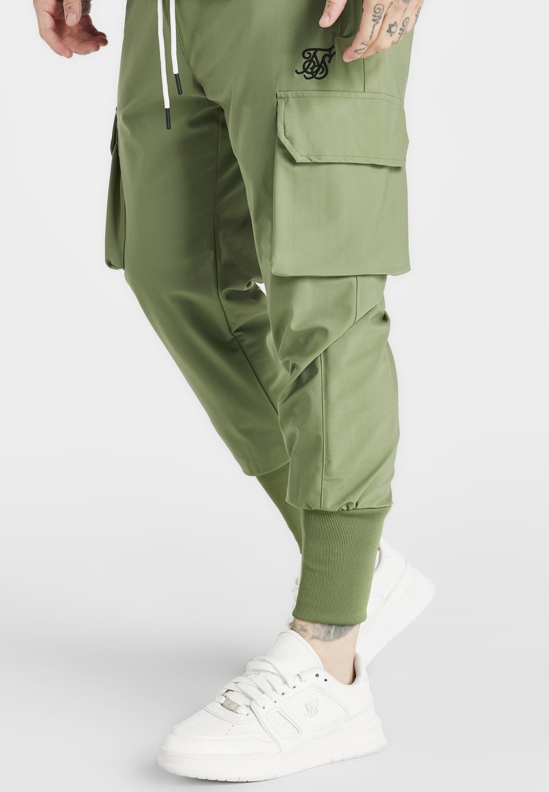 Grüne Cargo-Hosen mit mehreren Taschen, elastischen Bündchen und einem Taillenband mit Kordelzug. Kombiniert mit weißen Sneakers mit strukturiertem Design.